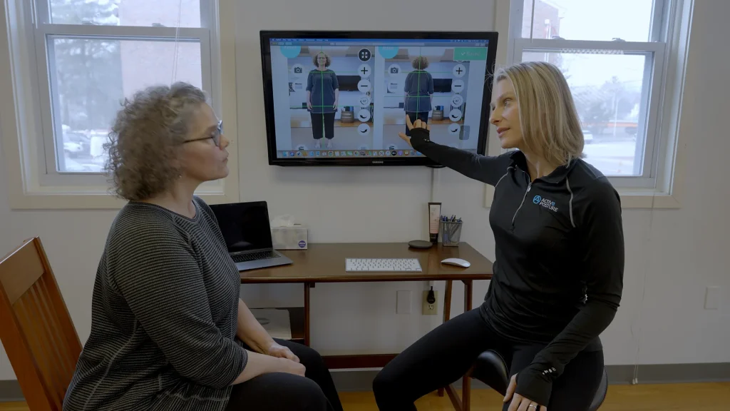 Posture therapist pointing out alignment findings on a wall-mounted screen to a seated client