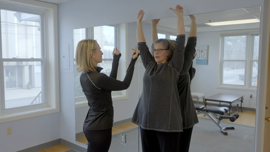 Therapist guiding a client through a standing overhead arm stretch beside the studio windows